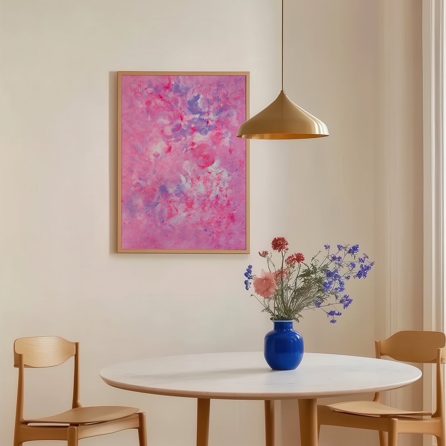 An abstract pink and purple painting in a light wood frame hangs on a beige wall above a round white dining table with two wooden chairs and a blue vase of flowers.
