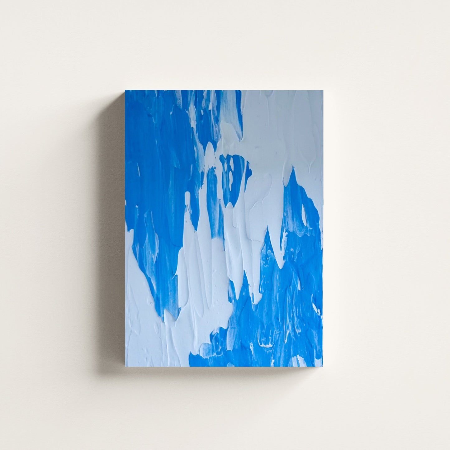 An abstract canvas painting featuring textured, thick brushstrokes of vibrant blue and light blue on a white background, shown hanging on a neutral off-white wall.
