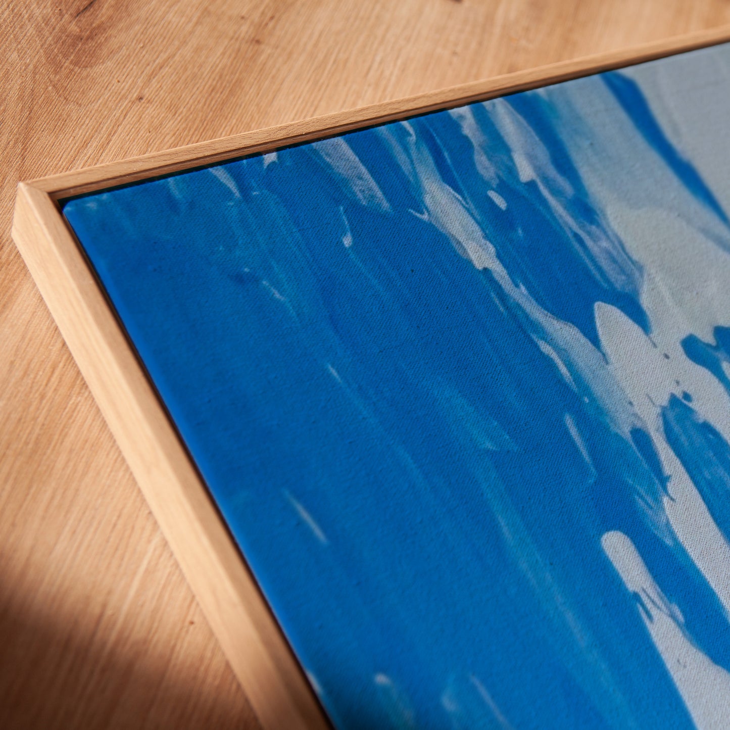 A close-up of a framed abstract painting featuring textured blue and white brushstrokes, set in a light natural wood frame on a wooden floor.