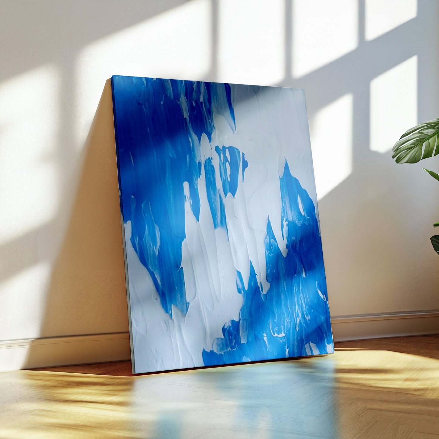 An abstract canvas painting with vibrant blue and white brushstrokes, leaning against a beige wall on a wooden floor next to a potted green plant in a sunlit room.