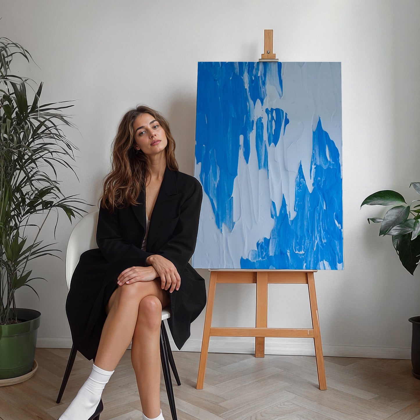A woman in a black blazer and white socks sits on a white chair next to a wooden easel displaying an abstract blue and white painting, with potted plants on either side in a bright room.