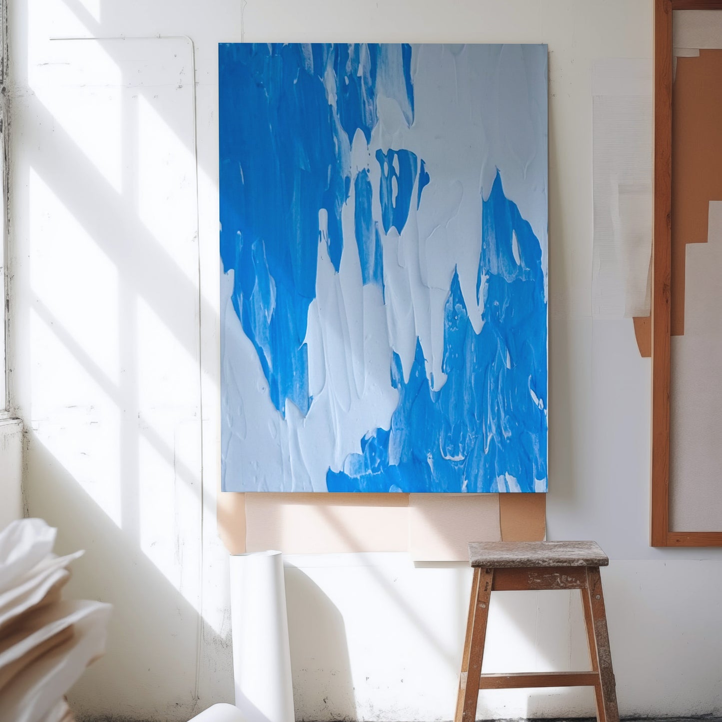 An abstract canvas painting with bold blue and white brushstrokes hanging on a white wall in a sunlit studio, with a wooden stool below and rolled paper on the floor nearby.