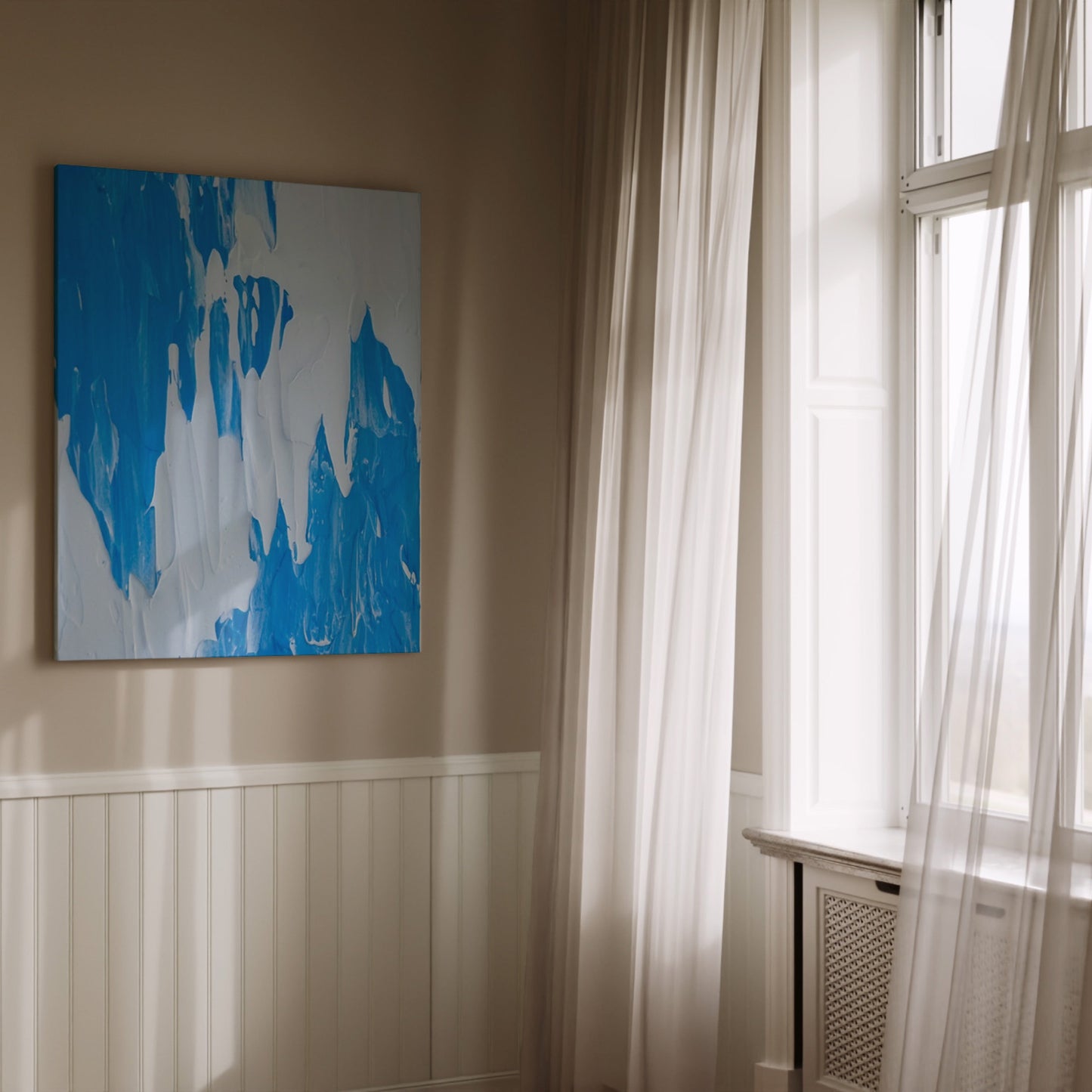 An abstract canvas painting with blue and white brushstrokes hanging on a beige wall in a sunlit room with white curtains and window trim.