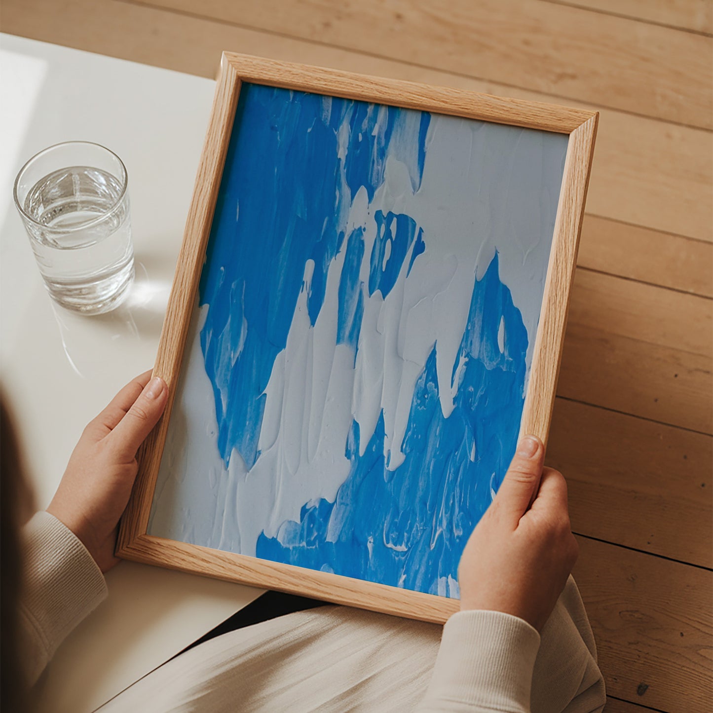 A person holds a square framed abstract painting with blue and white paint strokes in a light wood frame, photographed from above on a white table with a glass of water and a wooden floor.