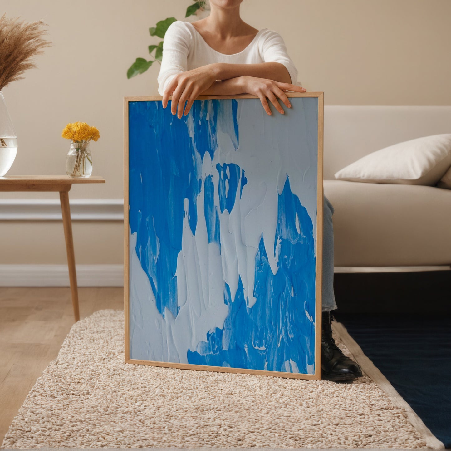 A person in a white top holds a large framed abstract painting featuring bold blue and white brushstrokes, set in a modern living room with a beige sofa, side table, and rug.