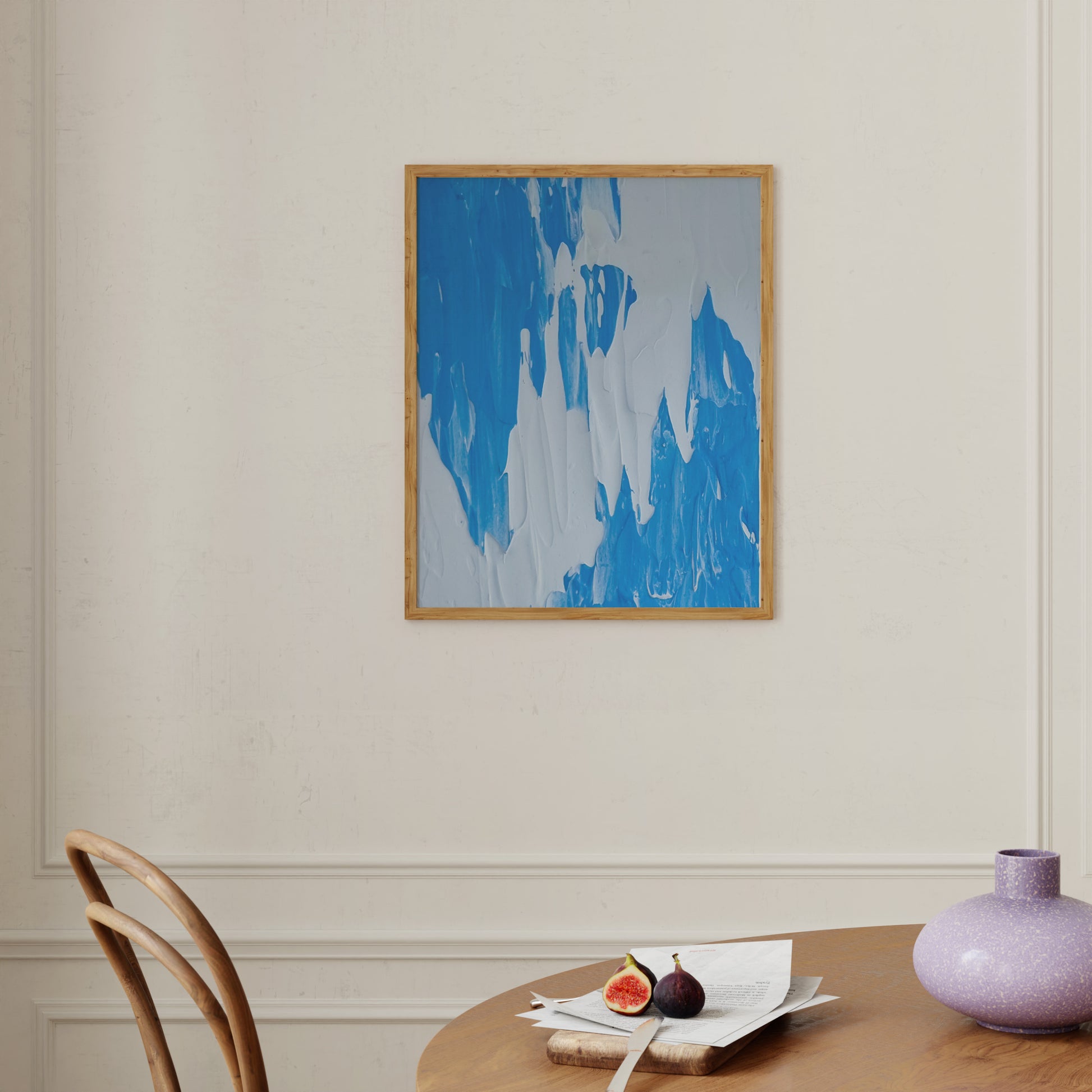A framed abstract painting with blue and white brushstrokes hangs on a cream-colored wall above a wooden dining table with a chair, papers, and a purple vase.