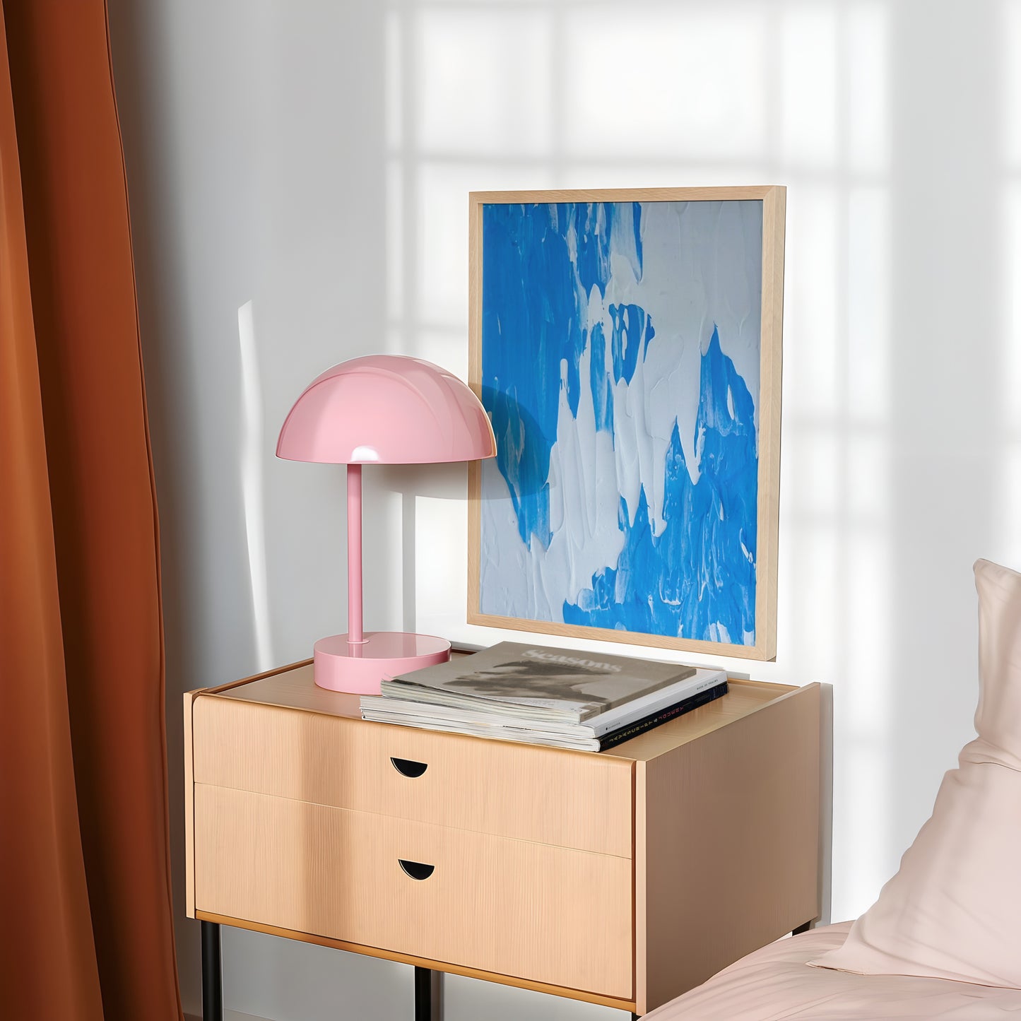 A light wood bedside table in a styled bedroom setting, holding a pink dome lamp and books, with a framed blue abstract print leaning against the wall.