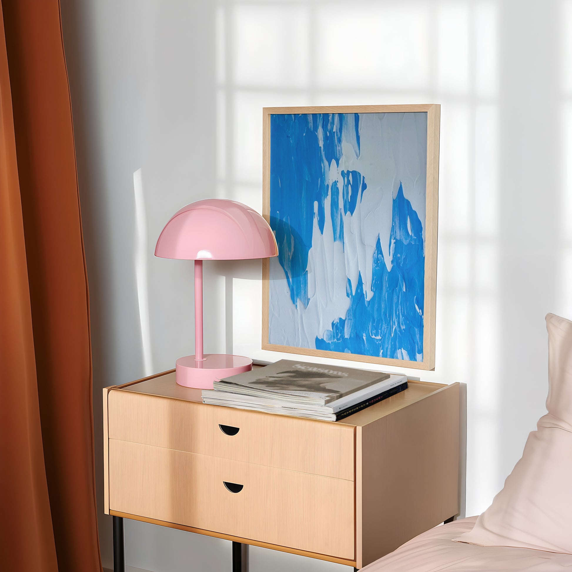 A light wood bedside table in a styled bedroom setting, holding a pink dome lamp and books, with a framed blue abstract print leaning against the wall.