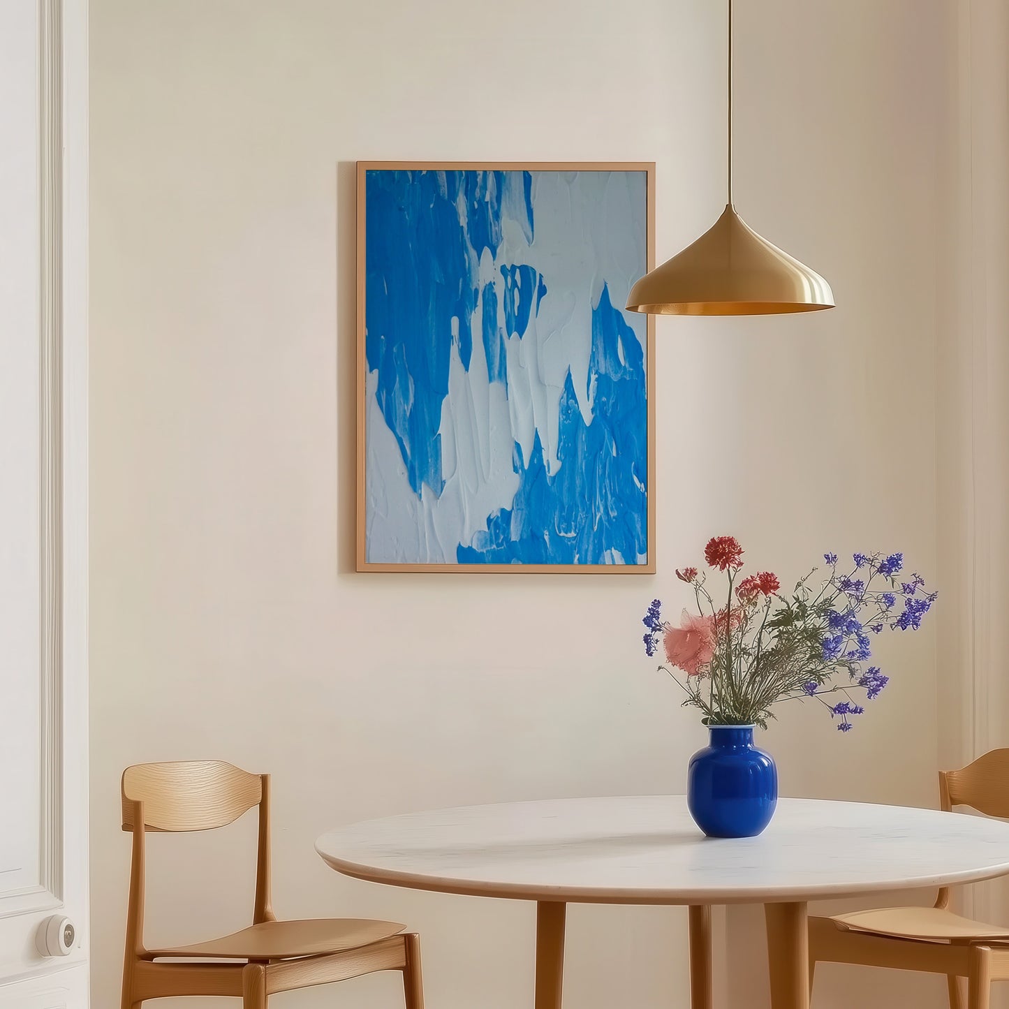 A light wood bedside table in a styled bedroom setting, holding a pink dome lamp and books, with a framed blue abstract print leaning against the wall.