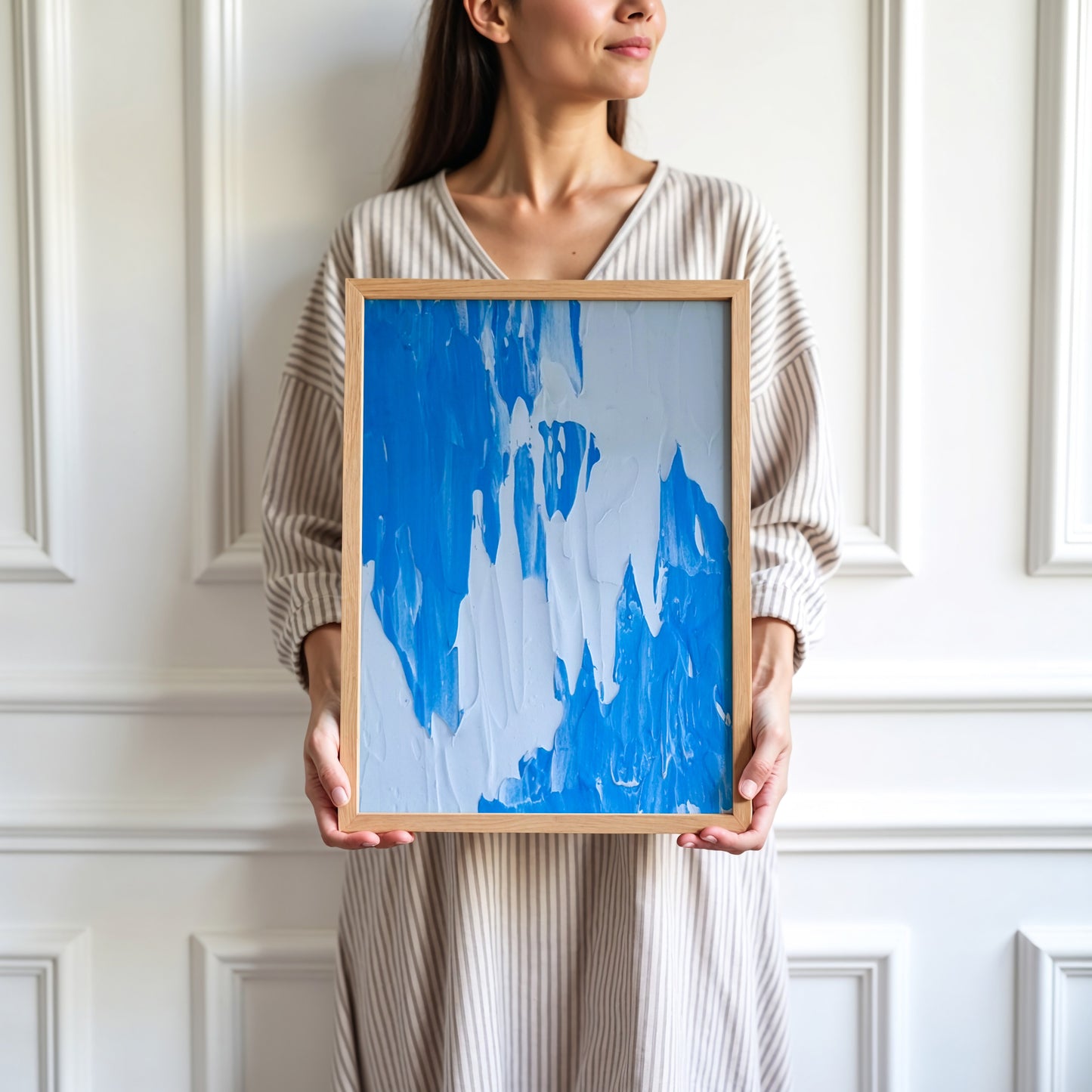 A woman in a beige striped V-neck dress holds a framed abstract painting featuring bold blue brushstrokes on a light background, set against a white paneled wall.