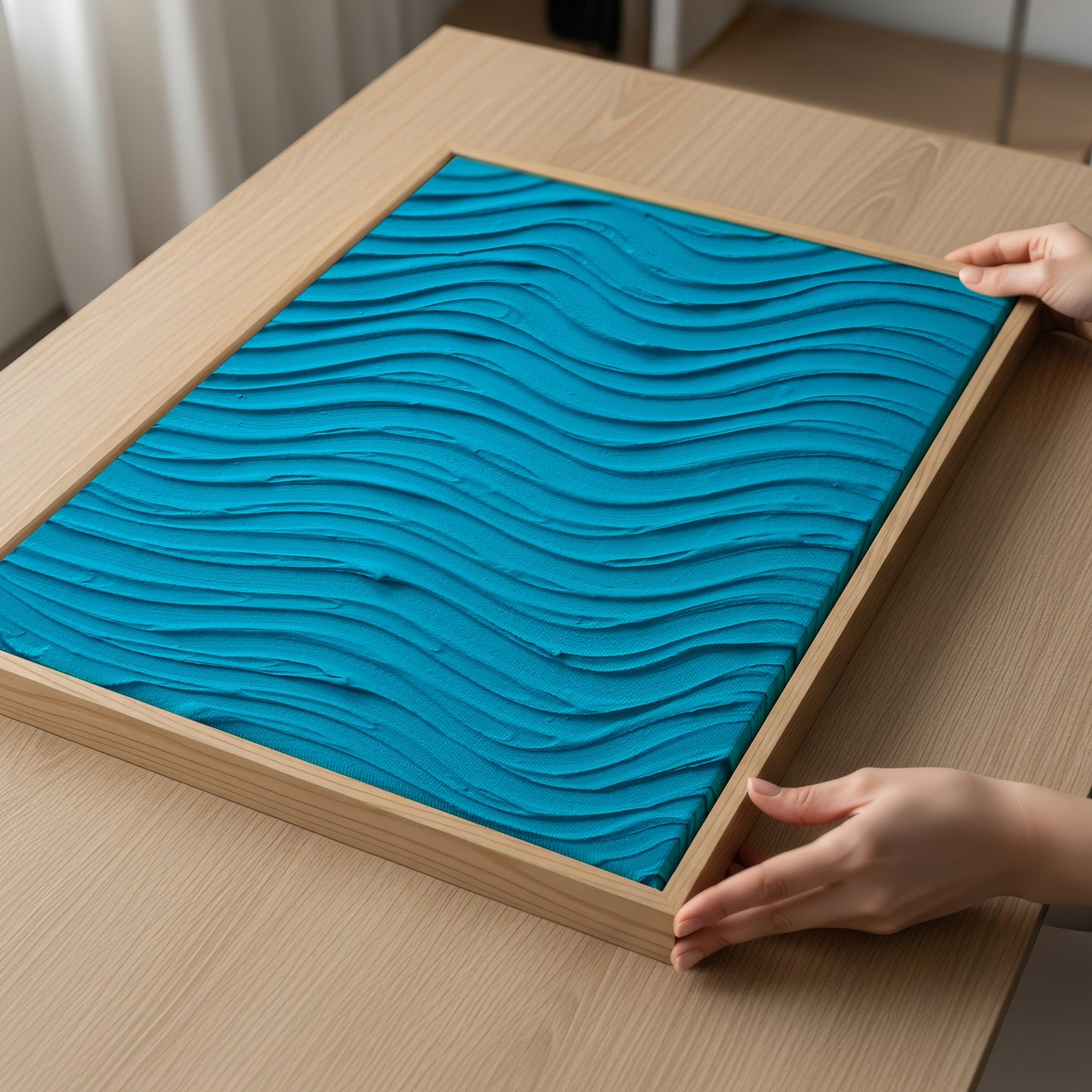 A square framed art print featuring a 3D wavy pattern in bright teal blue, displayed on a light wood table with hands adjusting the frame.