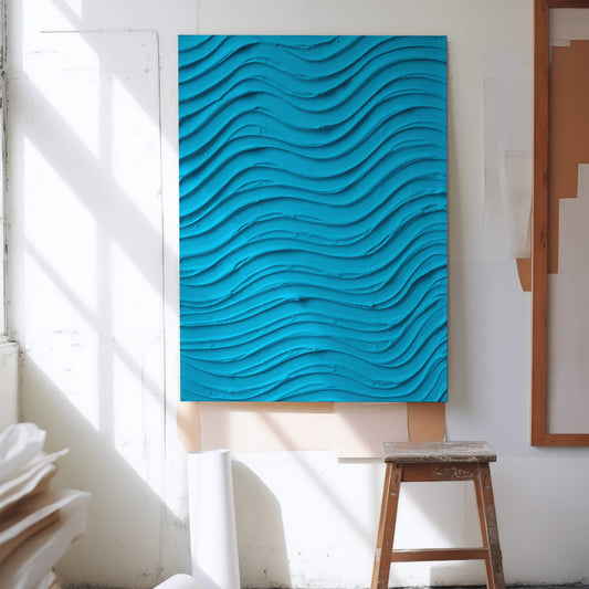 A large vertical wall art piece with a bright turquoise blue wavy, 3D wave pattern, displayed in a sunlit studio or gallery space next to a wooden stool.
