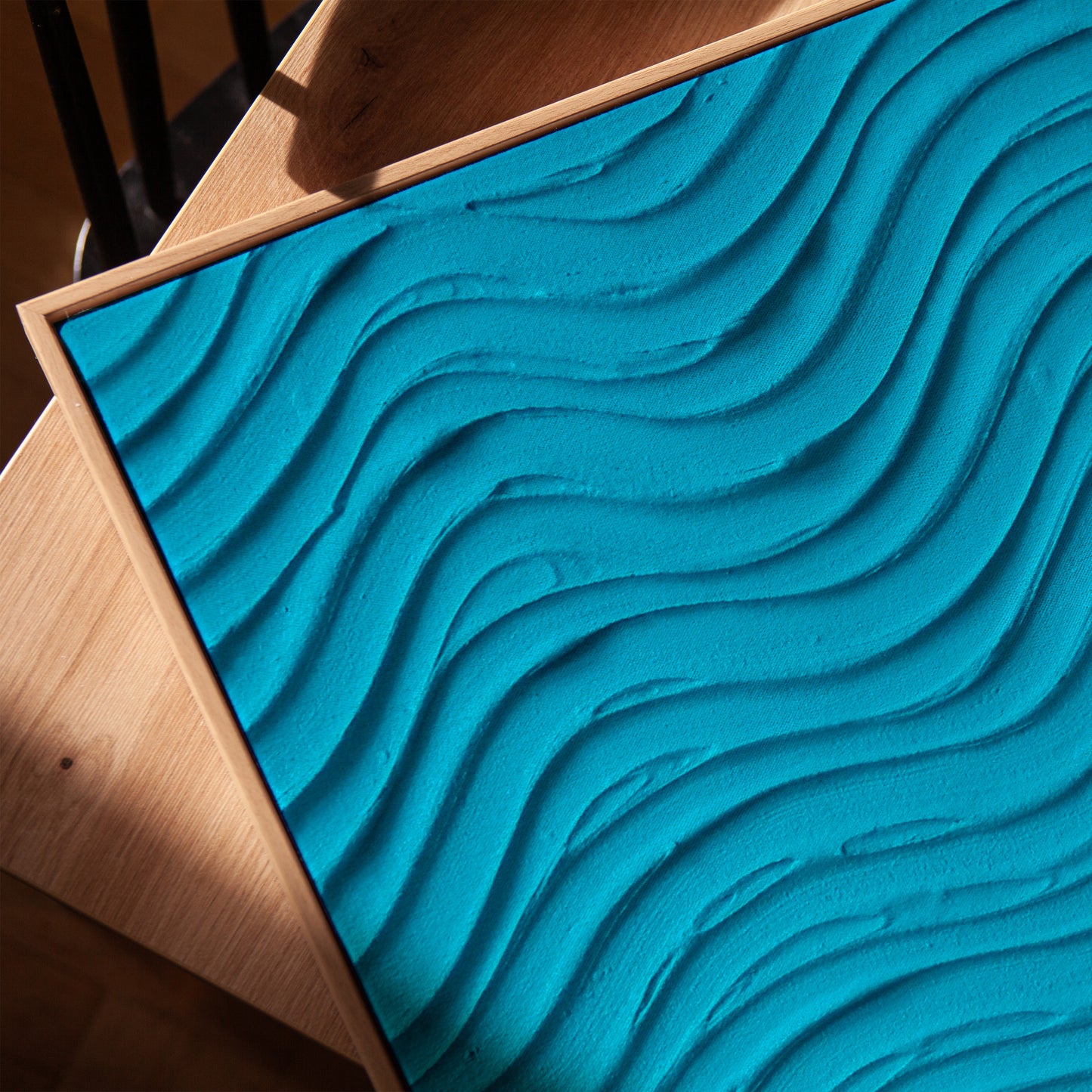 A square turquoise blue textured panel with raised wavy lines, mounted on a light wood frame and shown at an angle on a wooden surface.