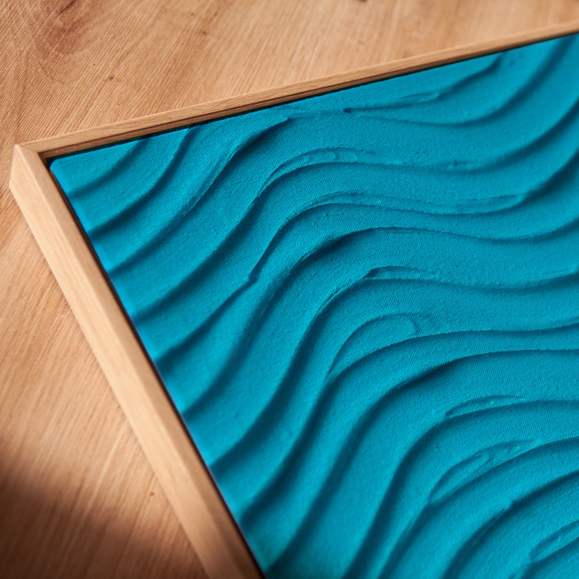 A square framed art piece featuring a bright turquoise blue textured surface with flowing, wave-like ridges, set in a light natural wood frame on a wooden tabletop.