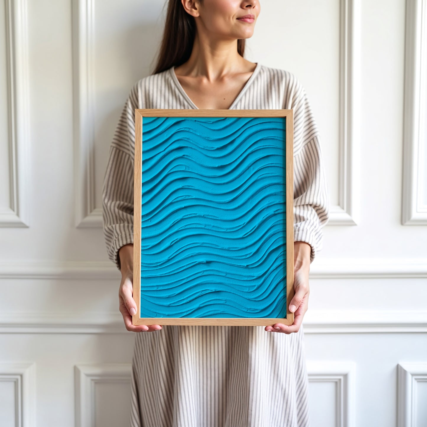 A woman in a beige striped dress holds a square framed art print featuring a bright turquoise blue 3D wavy texture, set against a white paneled wall.