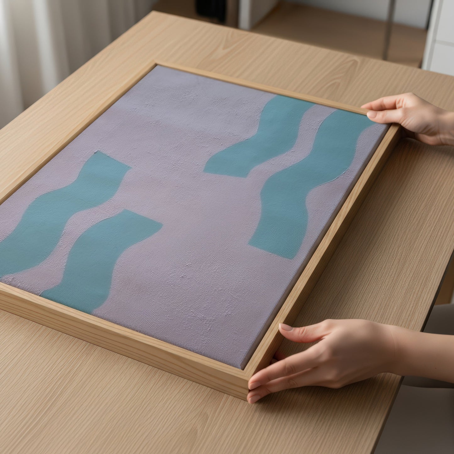 A framed abstract art print with wavy teal shapes on a muted lavender background, held by hands on a light wood table.
