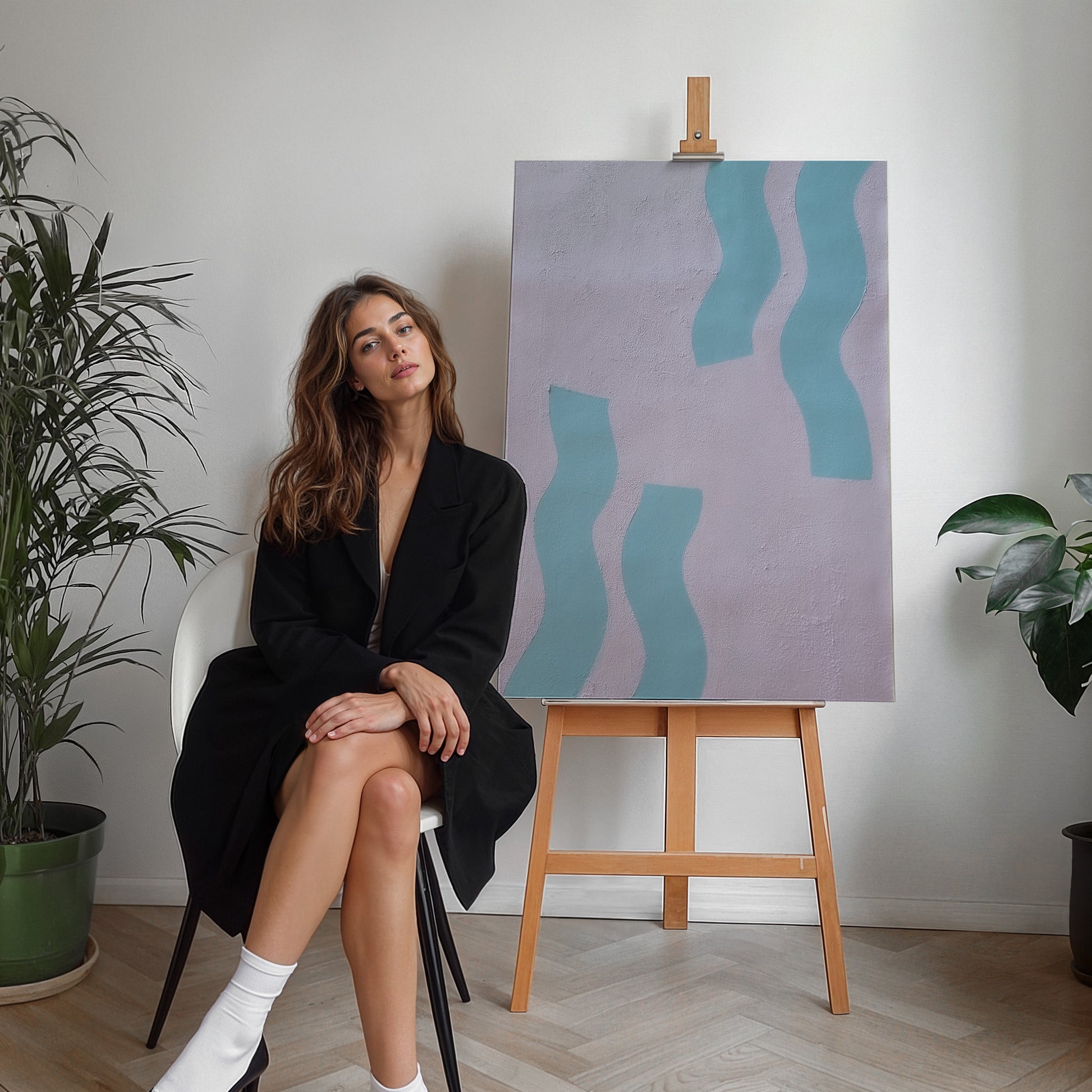 A woman in a black blazer sits on a white chair next to a wooden easel displaying an abstract painting with wavy light blue shapes on a lavender background. The room has light wood floors, white walls, and potted plants.