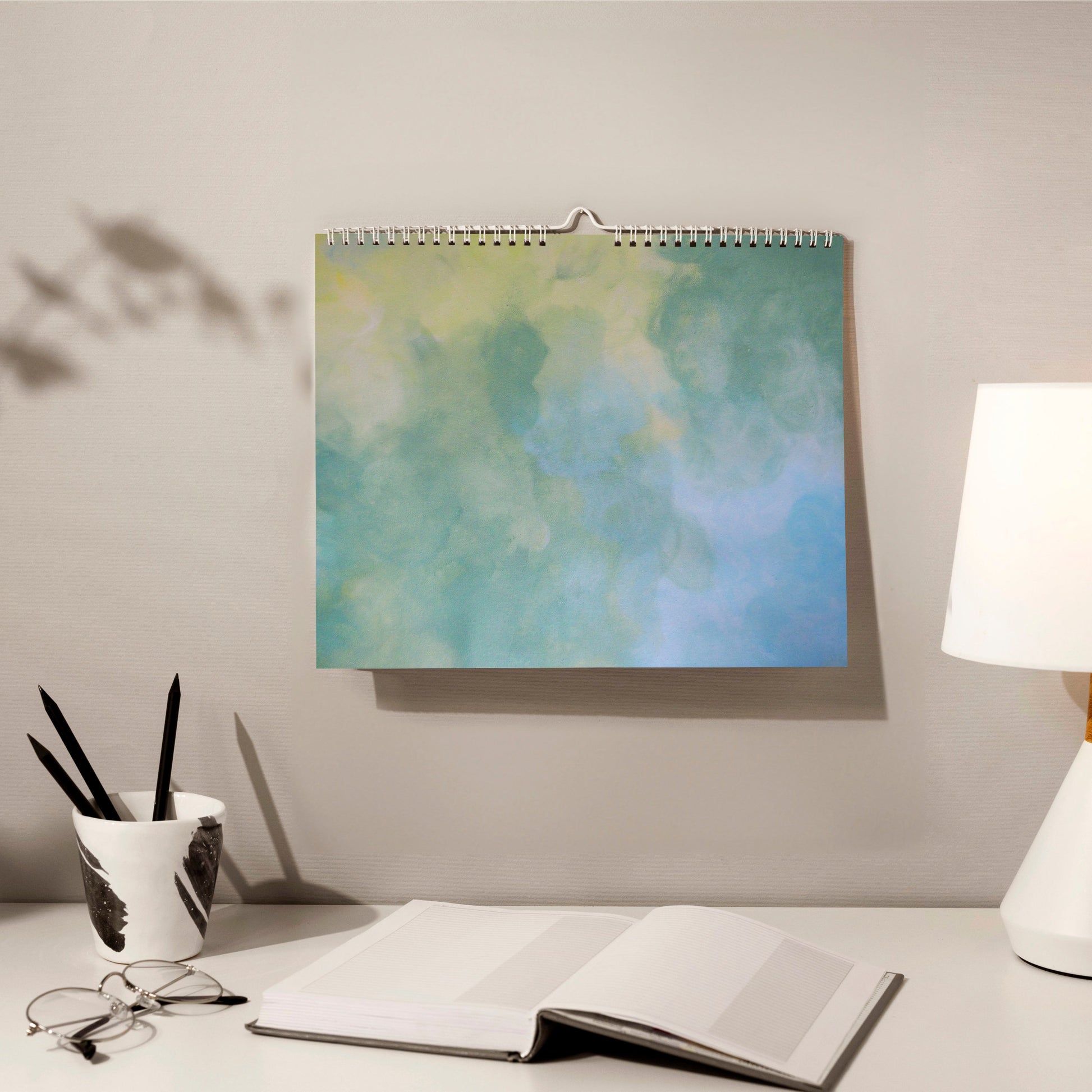 horizontal blue, green and yellow abstract art calendar by scandinavian artist hanging on a grey wall in a cozy home office with a book, pencils and glasses in front of it. 