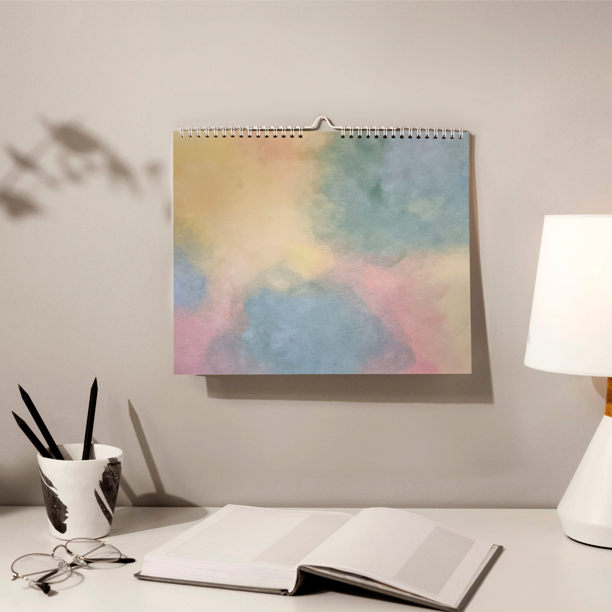 horizontal green, yellow and orange abstract art calendar by scandinavian artist hanging on a grey wall in a cozy home office with a book, pencils and glasses in front of it. 