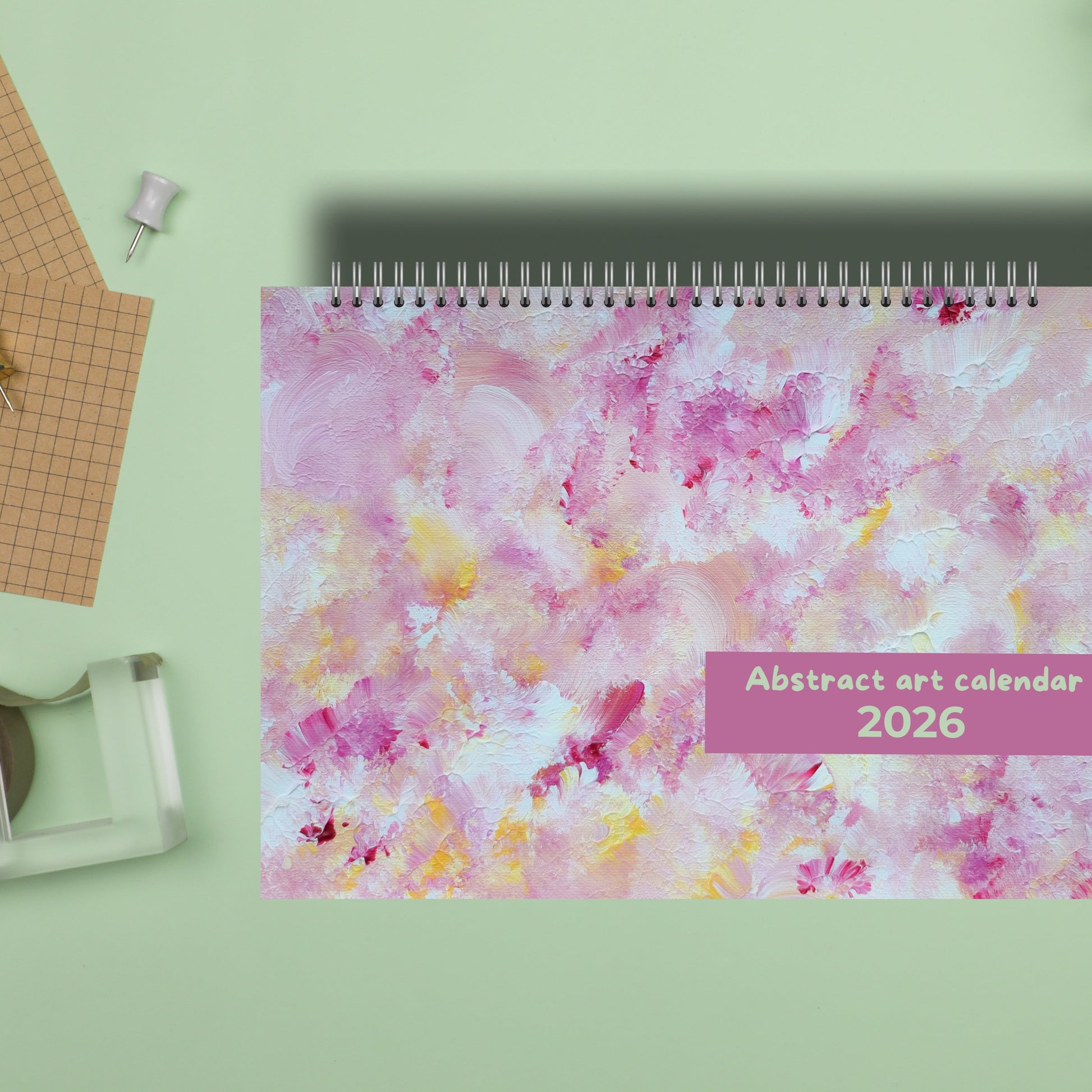 close up of horizontal pink abstract art wall calendar 2026 made by scandinavian artist Jeanette Haga, seen from above on a green desk with office accessorize and a green plant. 