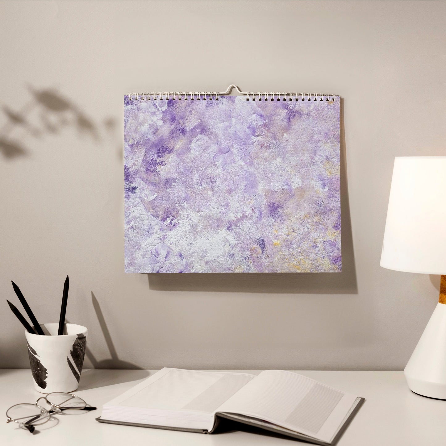 horizontal purple abstract art calendar by scandinavian artist hanging on a grey wall in a cozy home office with a book, pencils and glasses in front of it. 