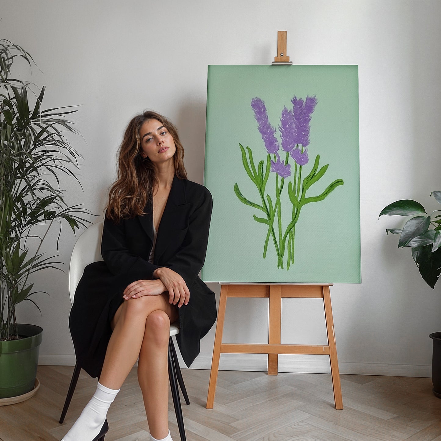 A woman in a black blazer sits on a white chair next to a wooden easel displaying a painting of purple lavender stems on a mint green background, in a bright room with potted plants.
