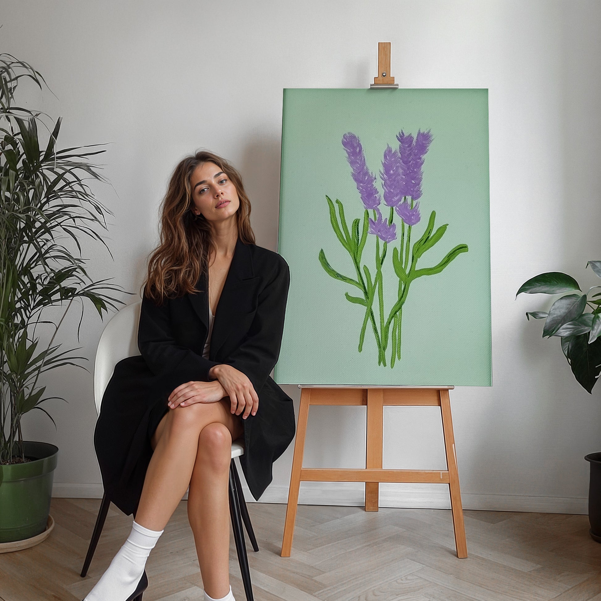 A woman in a black blazer sits on a white chair next to a wooden easel displaying a painting of purple lavender stems on a mint green background, in a bright room with potted plants.