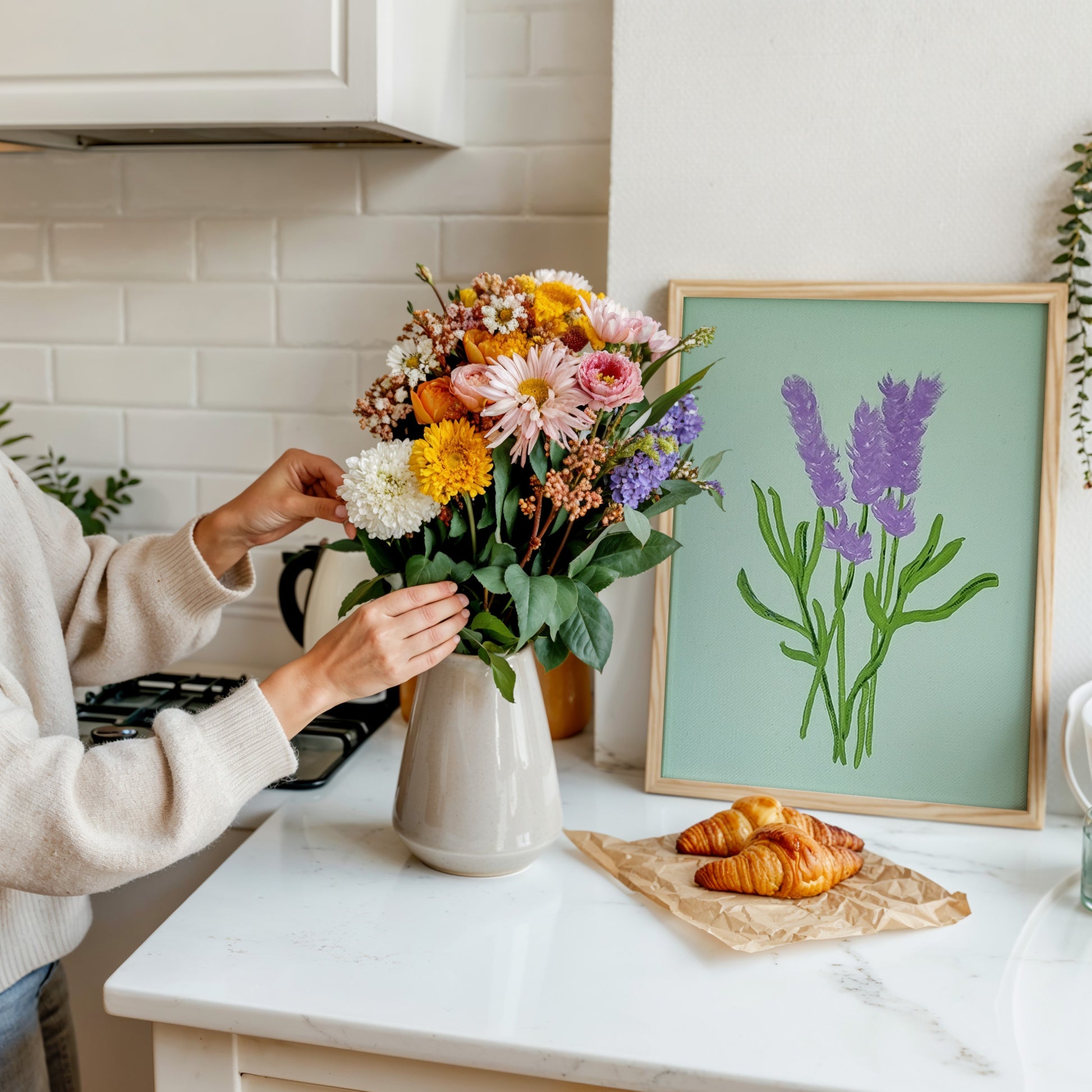 A person arranging a colorful bouquet of flowers in a white ceramic vase on a white marble kitchen counter, with a framed lavender illustration leaning against the wall behind them and croissants on a napkin nearby.