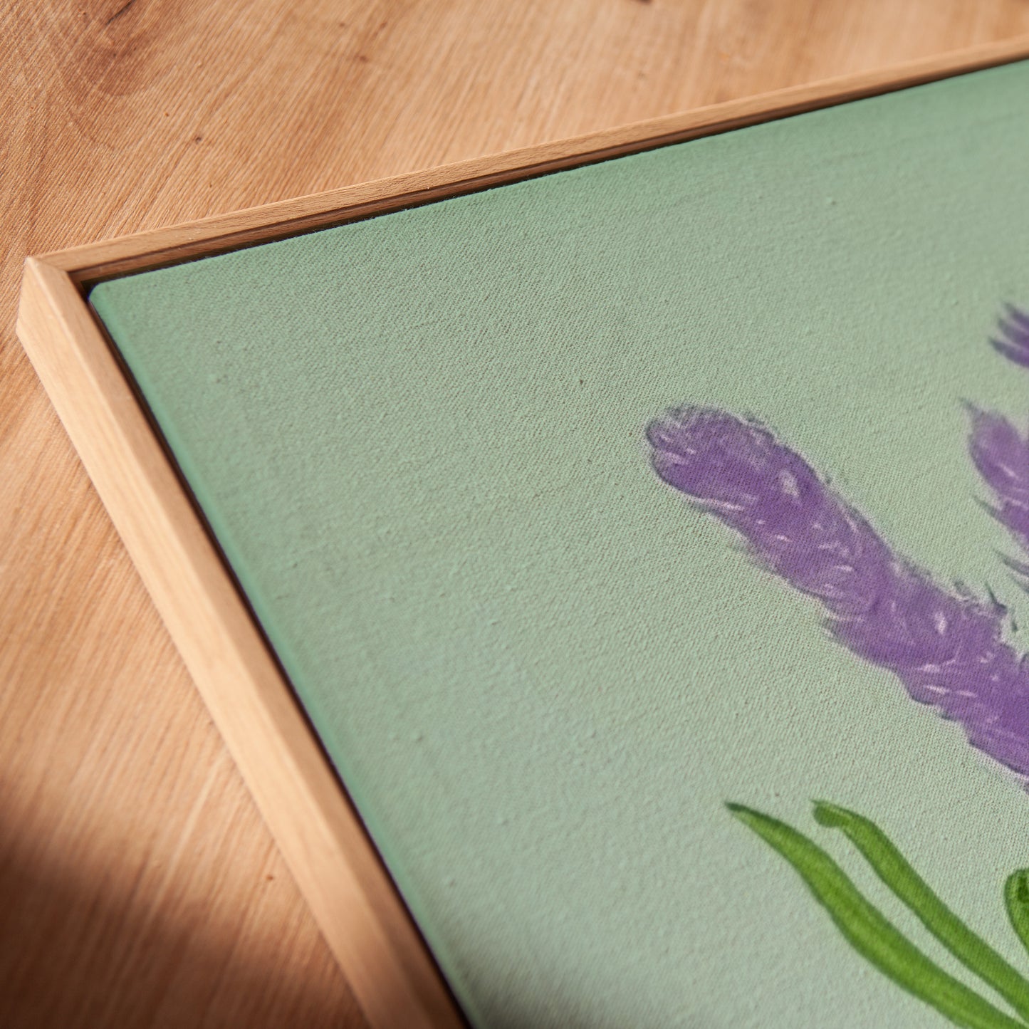 Close-up of a framed art print featuring a purple lavender flower and green leaves on a sage green background, set in a light wood frame on a wooden surface.