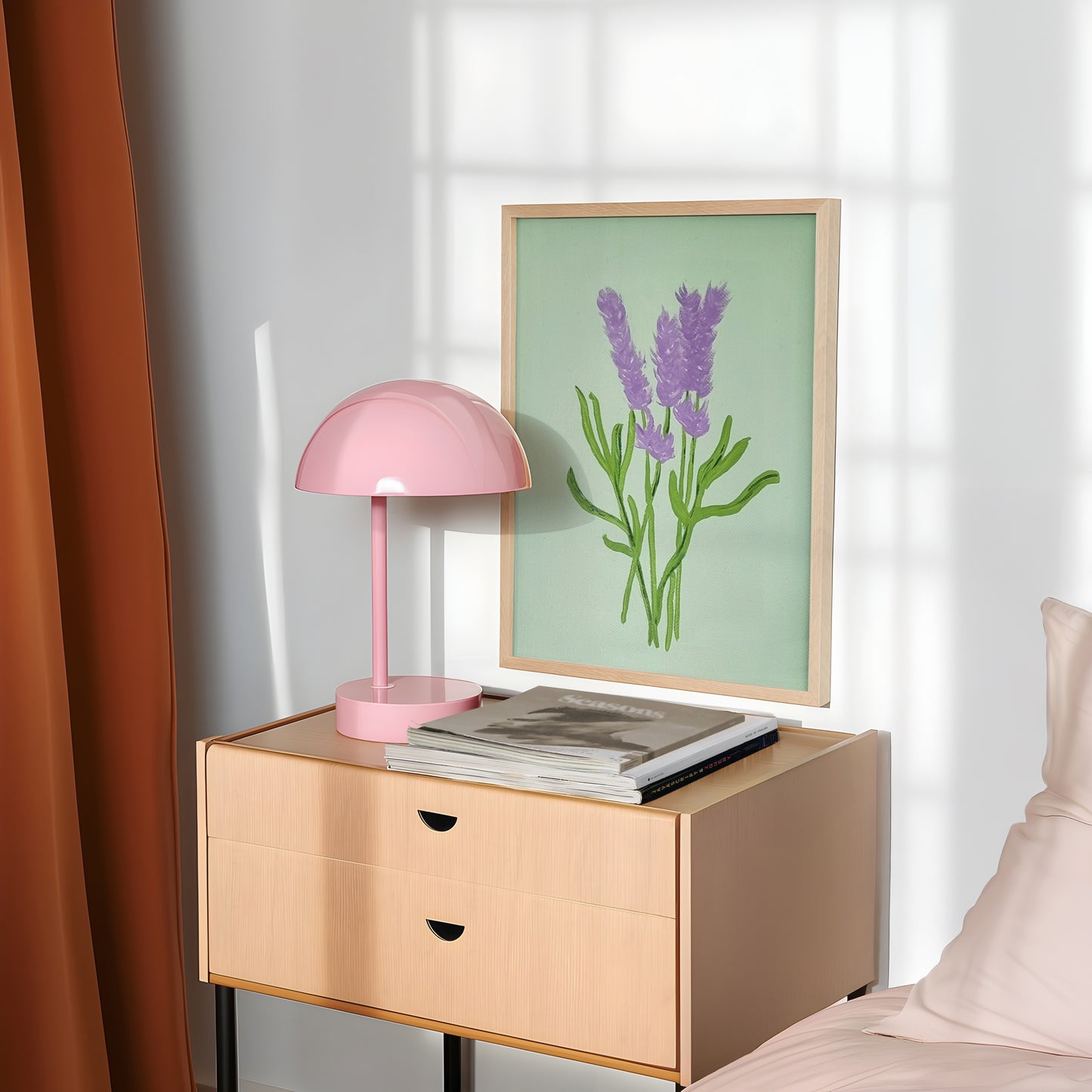 A light wood nightstand in a bedroom setting, styled with a pink dome lamp, a stack of magazines, and a framed botanical print of lavender flowers against a white wall with window shadows.