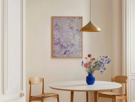 A framed abstract art print with purple and white brushstrokes hangs on a beige wall in a dining room setting above a round white table with wooden chairs and a blue vase of flowers.