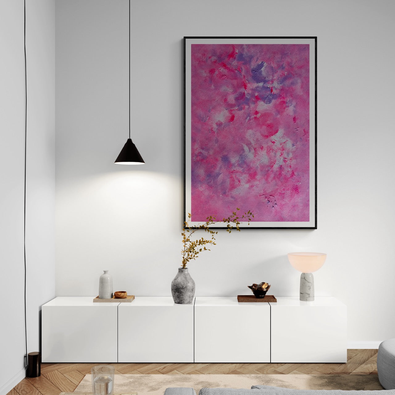 Pink, purple and white abstract art poster with a black frame hanging vertically on a white wall in a modern and minimalistic living room with white and black furniture and wood and herringbone patterned floor