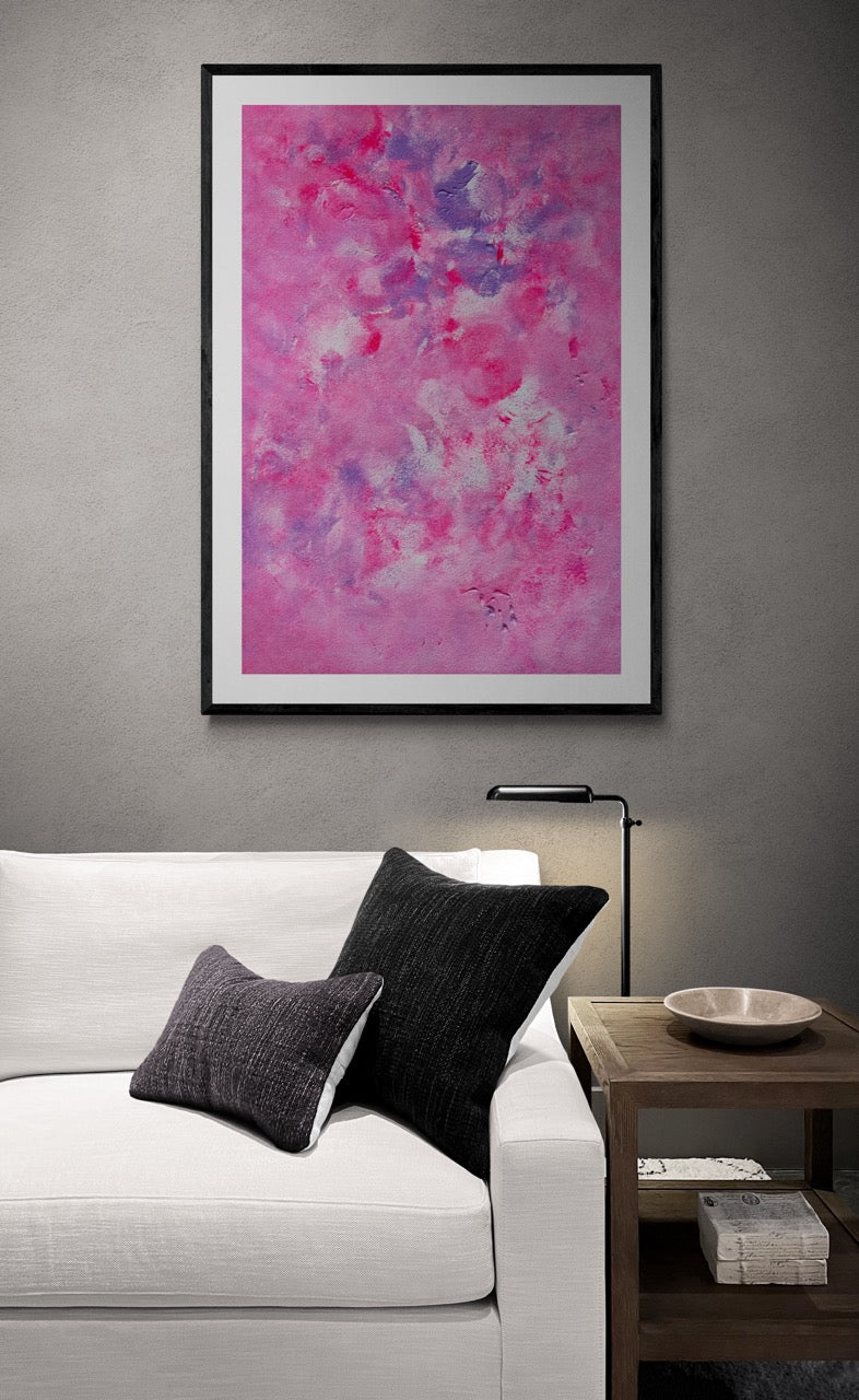 Pink, purple and white abstract art poster with a black frame hanging vertically on a grey wall behind a modern white sofa with black cushions on them. Next to the sofa is a walnut side table with a decor bowl on top.