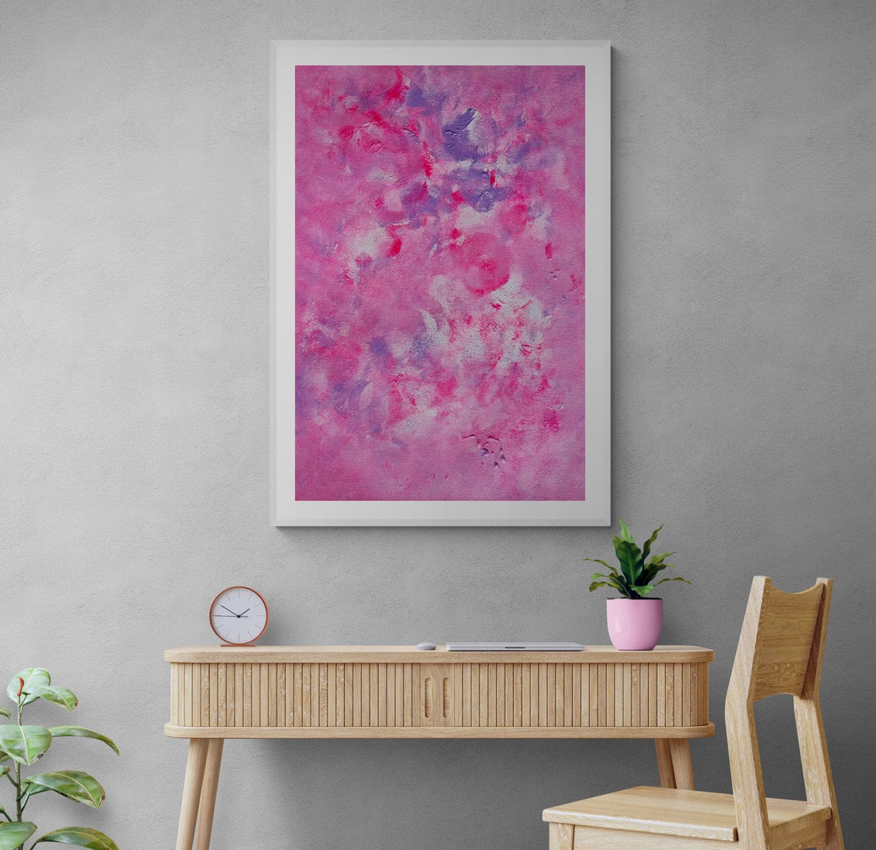 Pink, purple and white abstract art poster with a white frame hanging vertically on a light grey wall over a working desk. The room is an office and the furniture is of light oak. 